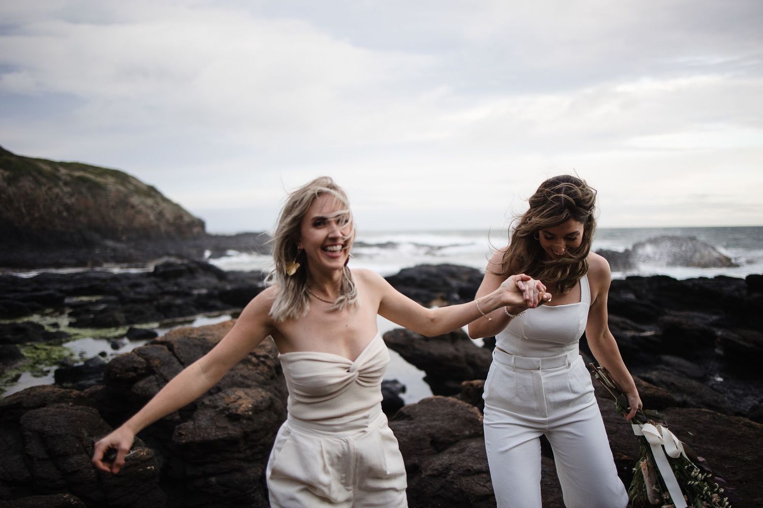 Two people in white outfits dance and laugh together on rocky coastal cliffs with ocean waves crashing behind them.