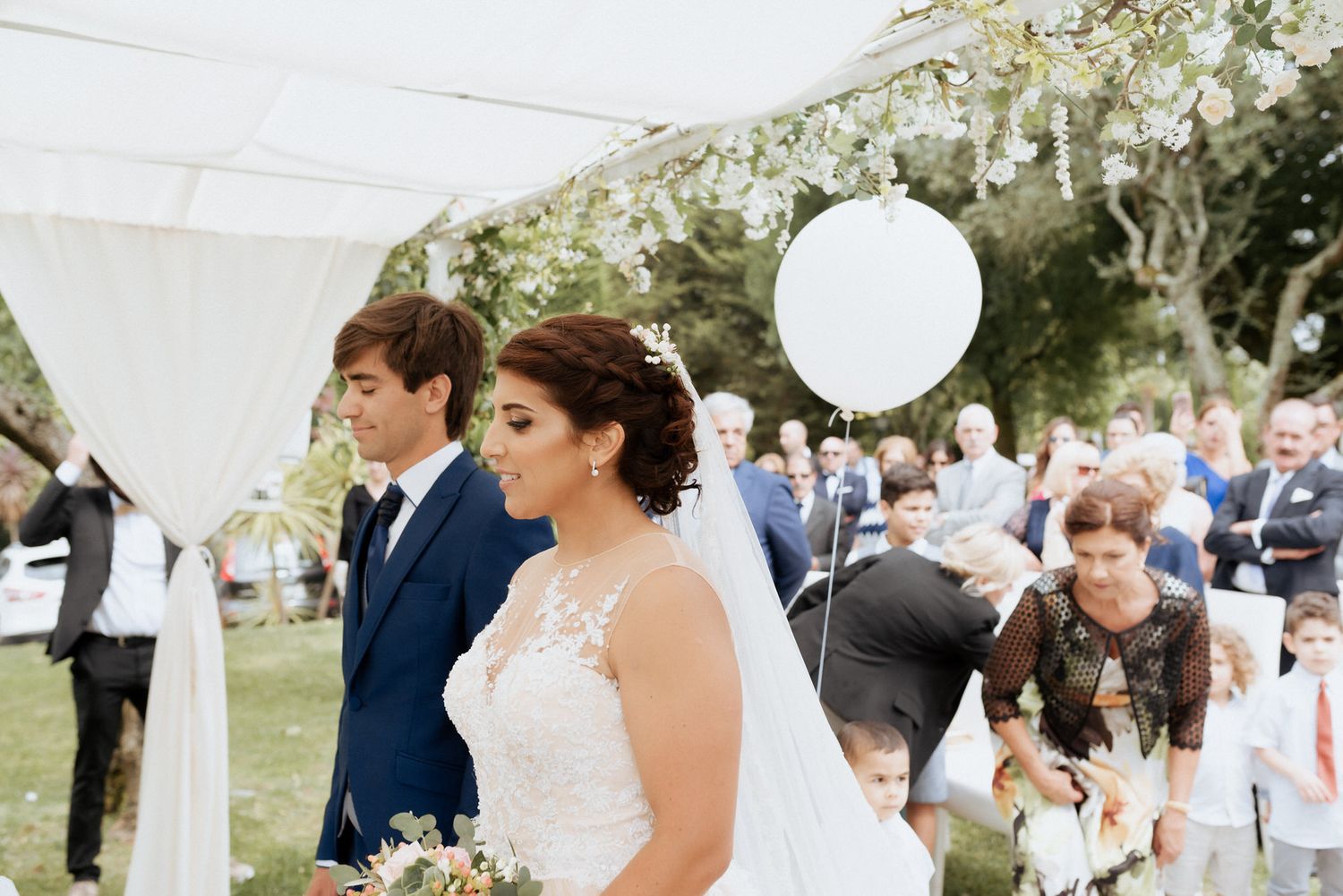 Classic outdoor wedding ceremony in a garden setting with white decor and guests gathered under trees.