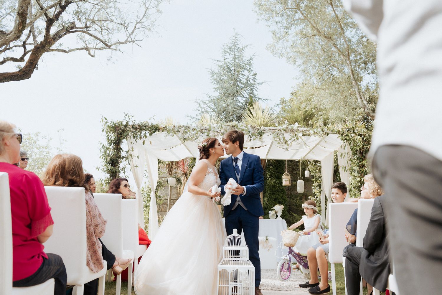 Romantic outdoor wedding ceremony under a white floral arch decorated with flowing white fabric on a sunny afternoon.