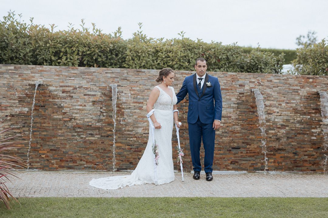 A couple in wedding attire stands before a rustic stone wall in an outdoor garden setting.