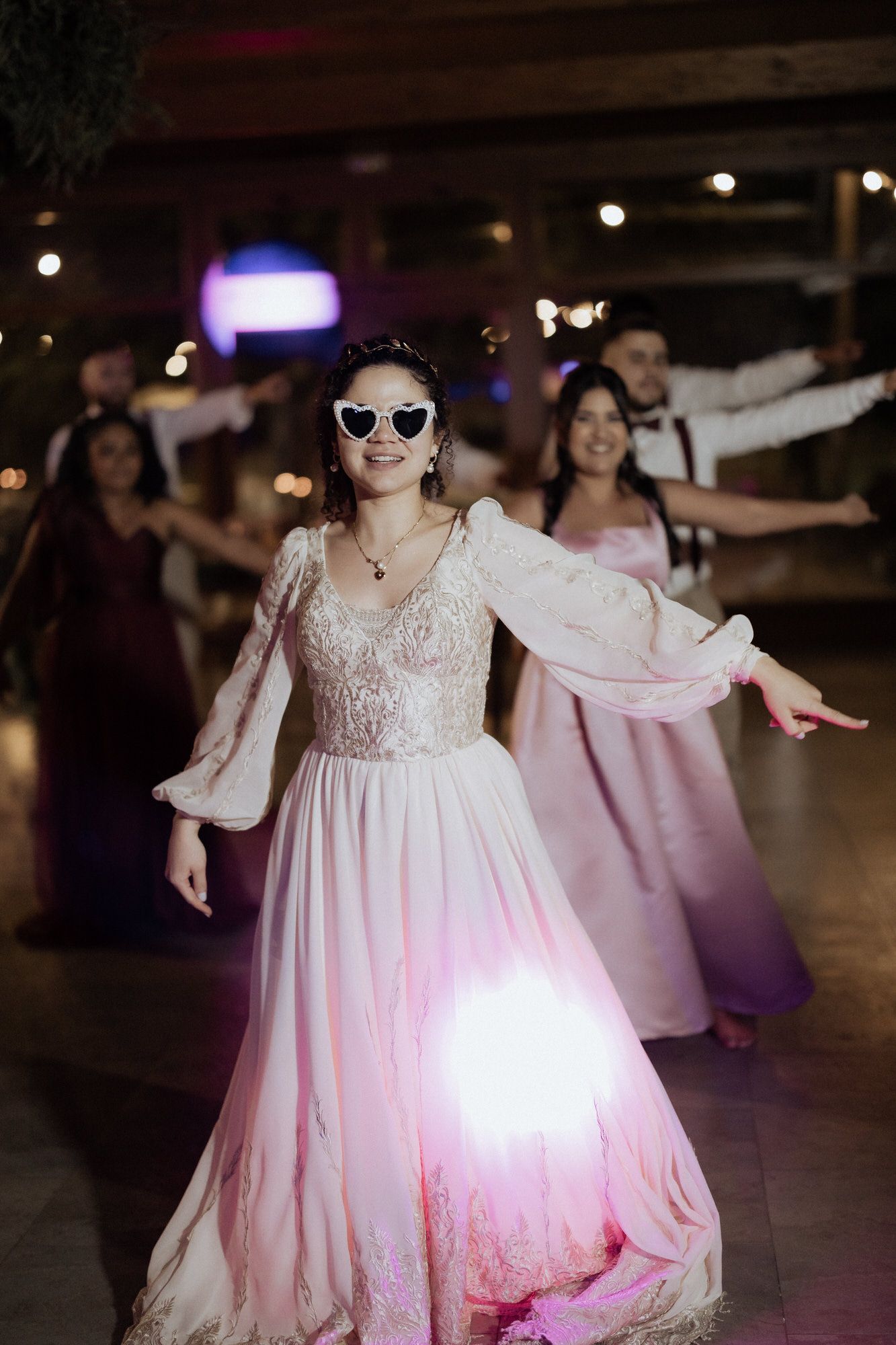 Woman in pink flowing gown and sunglasses dances at formal event with others visible in background.