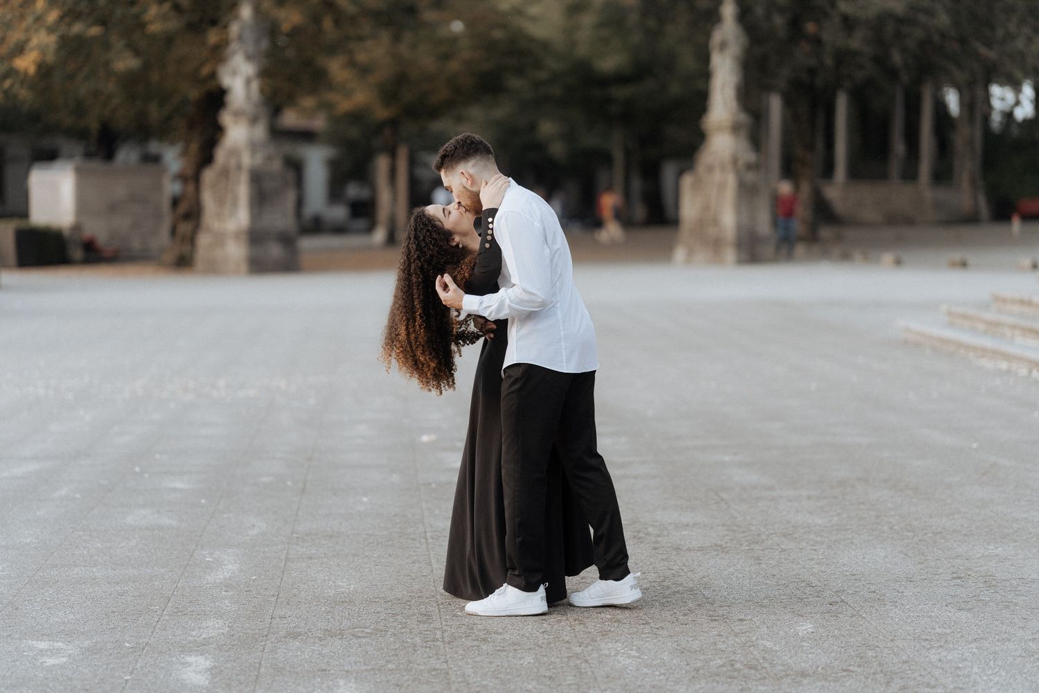 Um casal de mãos dadas enquanto caminhavam juntos em um pátio elegante com esculturas de pedra e árvores ao fundo.