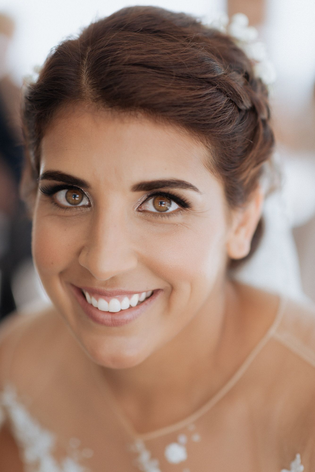 Close-up portrait showing radiant smile, elegant bridal makeup and upswept brown hair with blurred background.