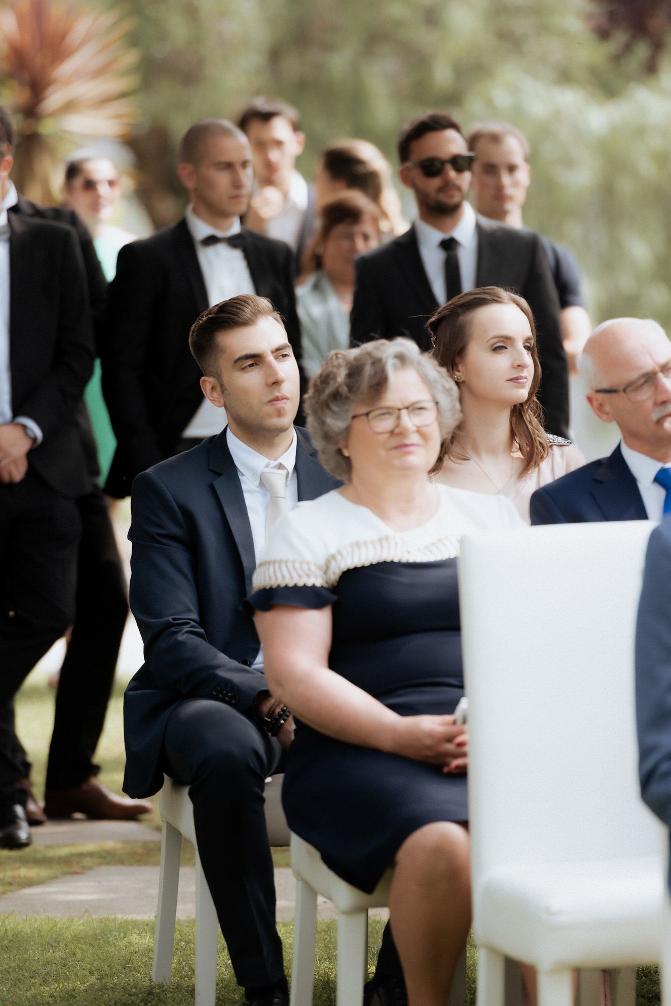 Wedding guests seated in white chairs during an outdoor ceremony in formal attire.