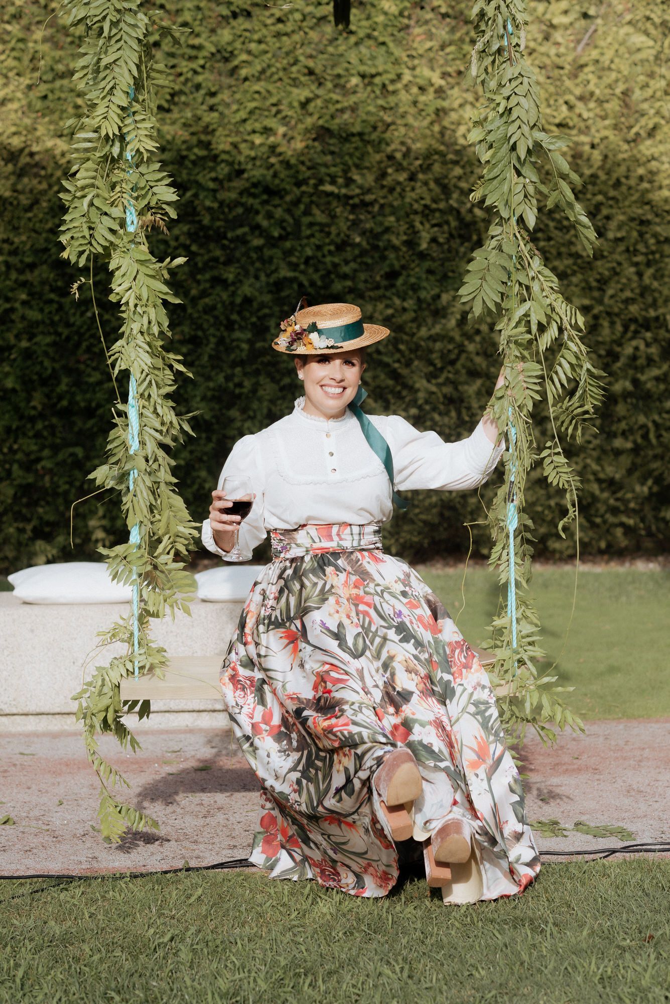 Woman in vintage-inspired floral dress and straw hat sits on garden swing decorated with hanging greenery.
