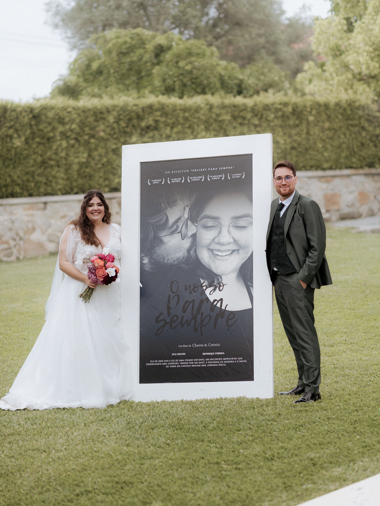A couple stands beside a large black and white movie-style poster on a manicured lawn with hedges in the background.