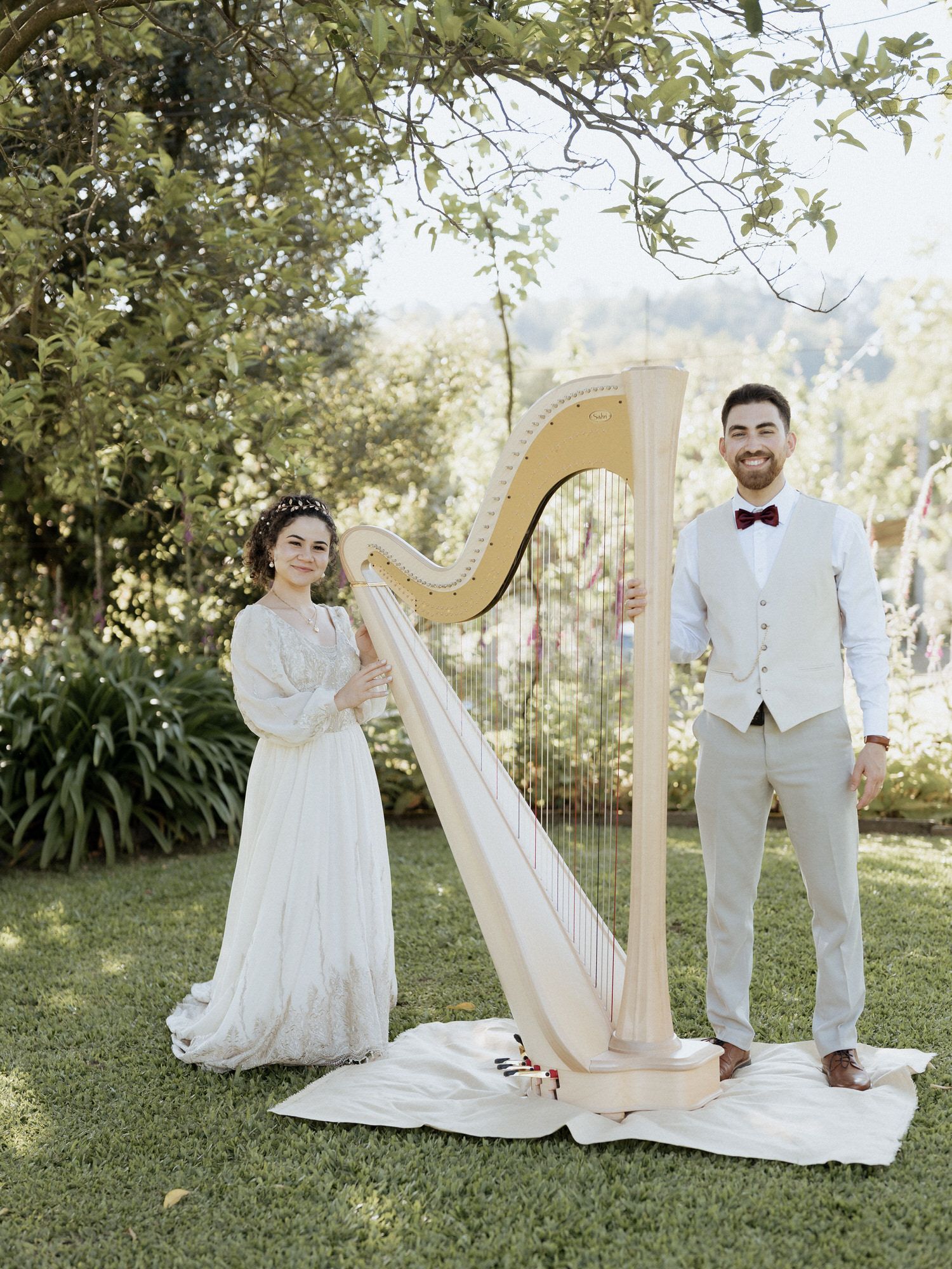 Couple poses outdoors at wedding with an elegant gold harp on a white cloth under tree branches in garden setting.