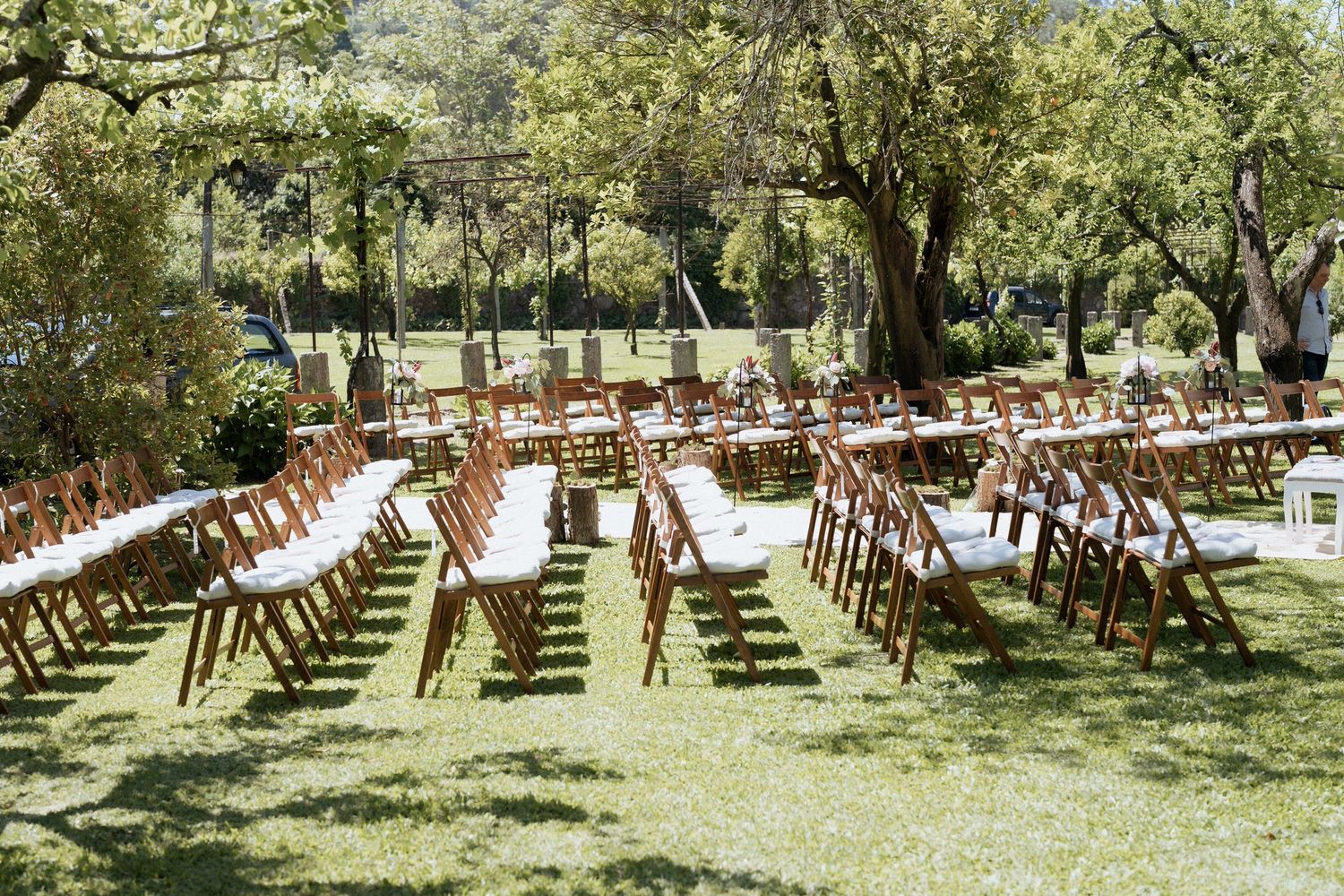 Wooden chairs arranged in rows on a lush green lawn for an outdoor garden wedding ceremony beneath shaded trees.