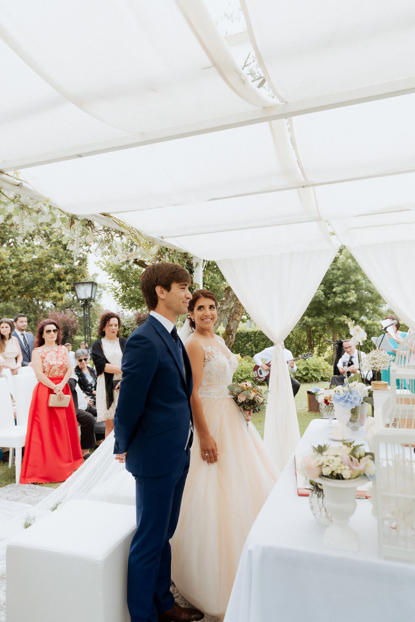 Wedding reception guests gather under elegant white canopy tent with floral arrangements and garden backdrop.