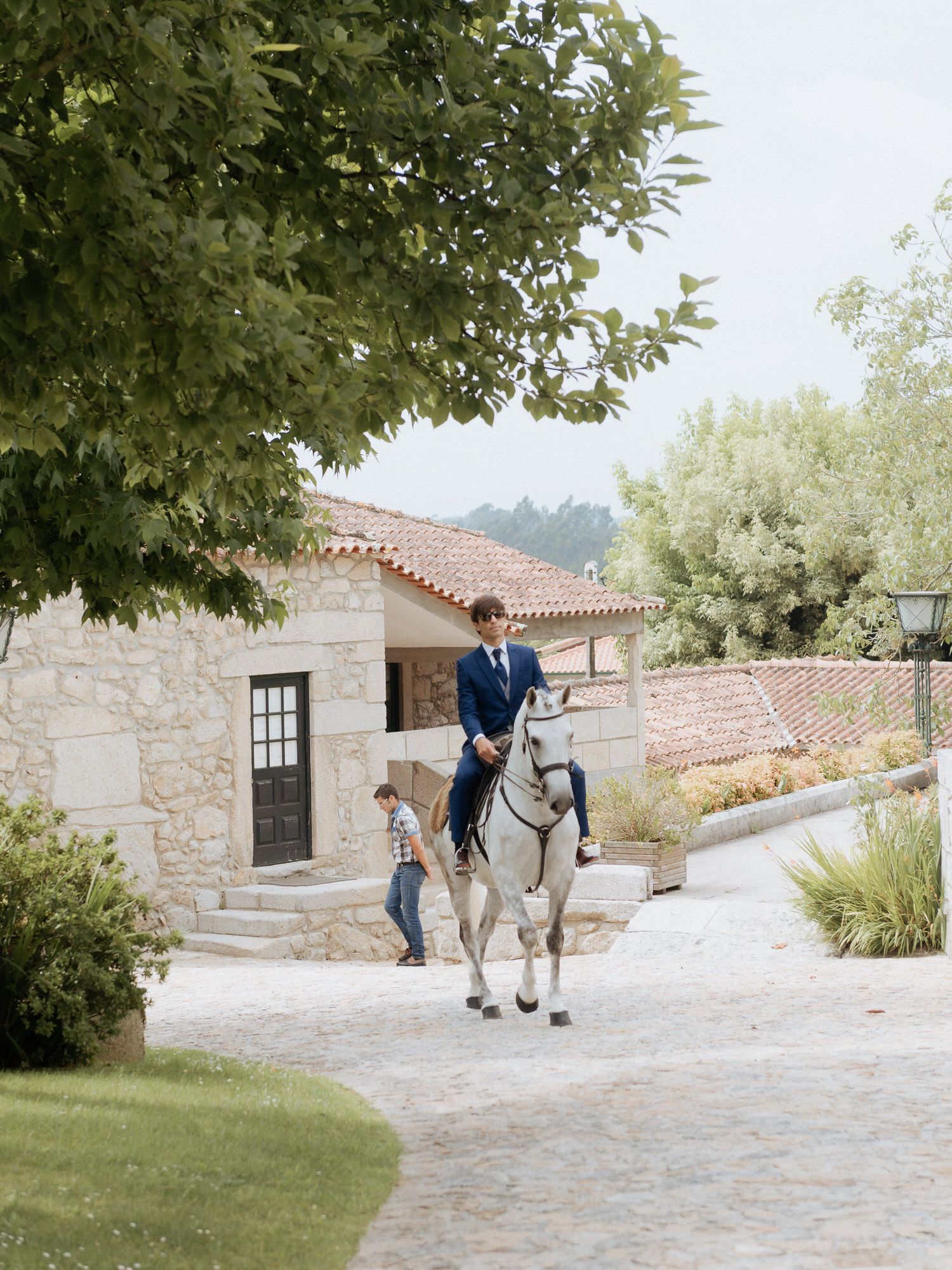 A person rides a white horse along a stone path beside a Mediterranean-style building with lush greenery and trees.