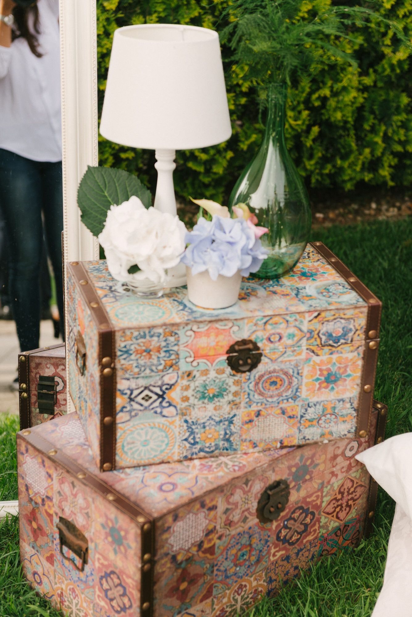 Vintage distressed storage chest with ornate floral designs serves as base for white table lamp on grass.