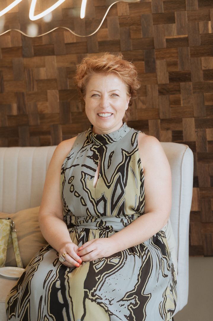 A person wearing a gray and black patterned sleeveless dress sits on a white chair against a textured brown wall.