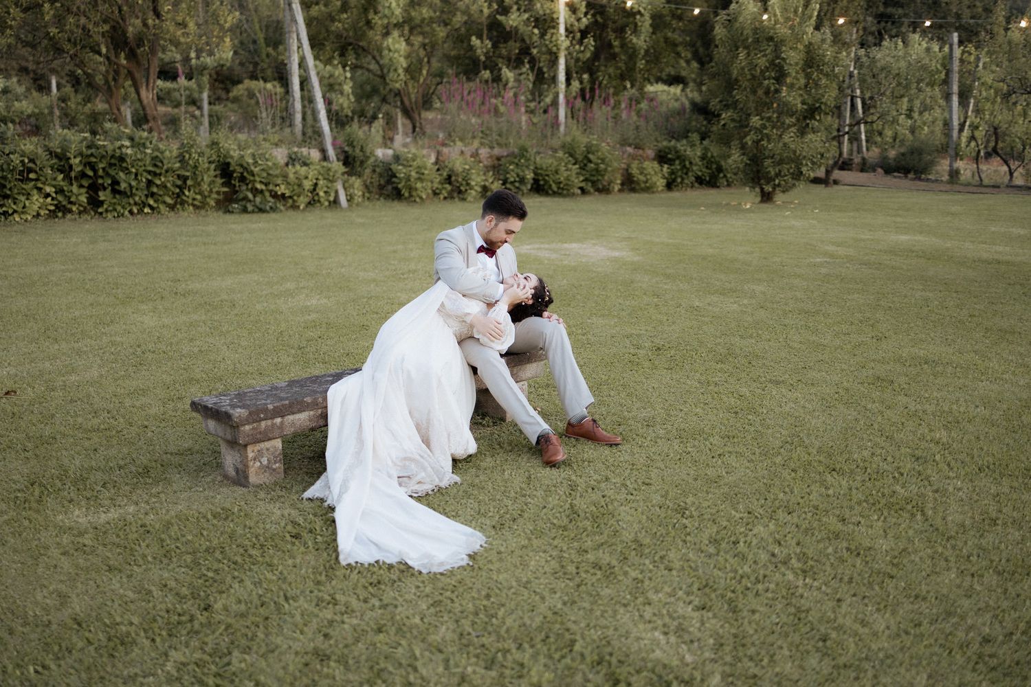 Two people in white wedding attire embrace while sitting on lush green grass in a garden setting.