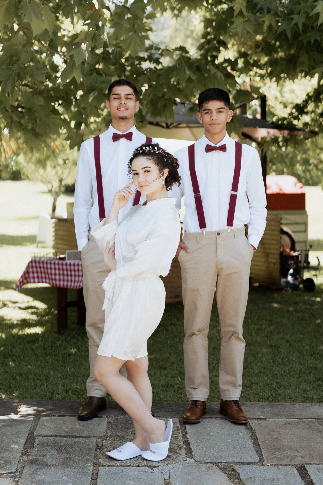 Three wedding party members pose outdoors in coordinated burgundy bow ties and suspenders with khaki pants.