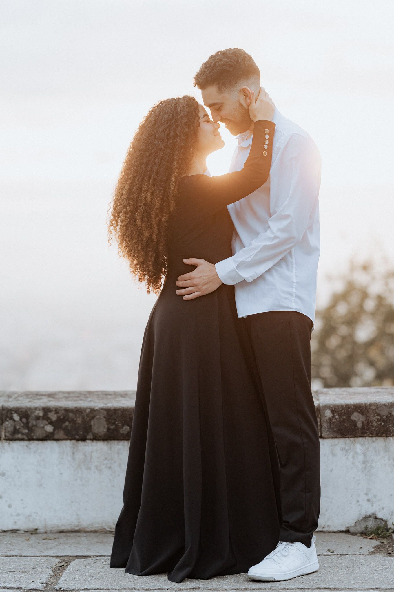 Um casal romântico se abraça durante a hora dourada, com uma mulher em um longo vestido preto e um homem em uma camisa branca.