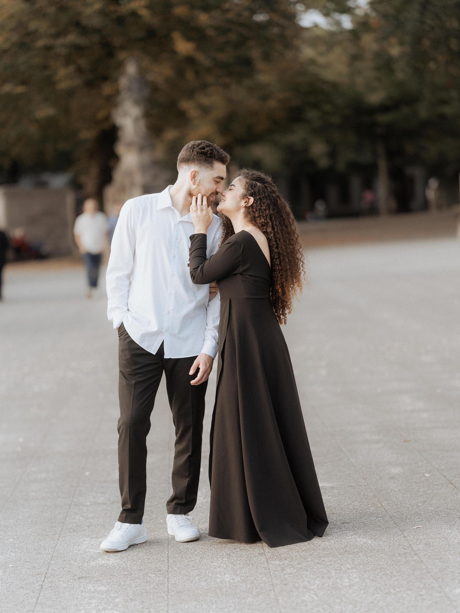 Um casal se abraça ternamente em um parque, ela usando um longo vestido marrom e ele uma camisa branca.
