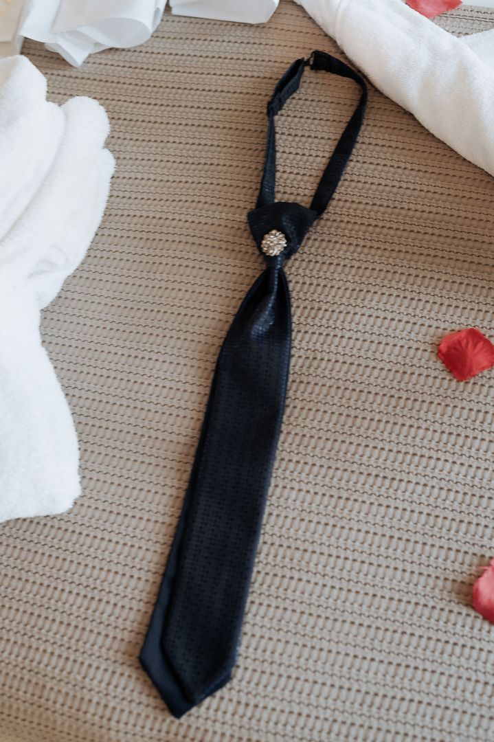 A black necktie with a decorative brooch pin lies on a beige textured surface with scattered rose petals and white fabric.