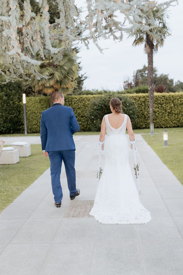 A bride in a white wedding gown and groom in navy suit walk along a palm-lined path at an outdoor venue.