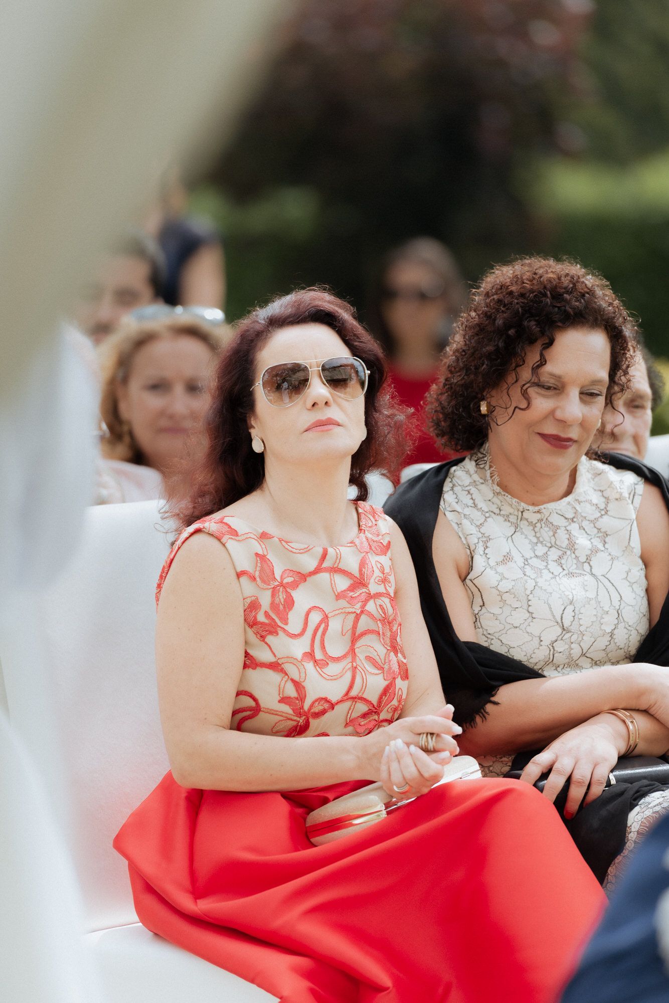 Wedding guests dressed in red and white dresses smile while seated outdoors at a summer ceremony.