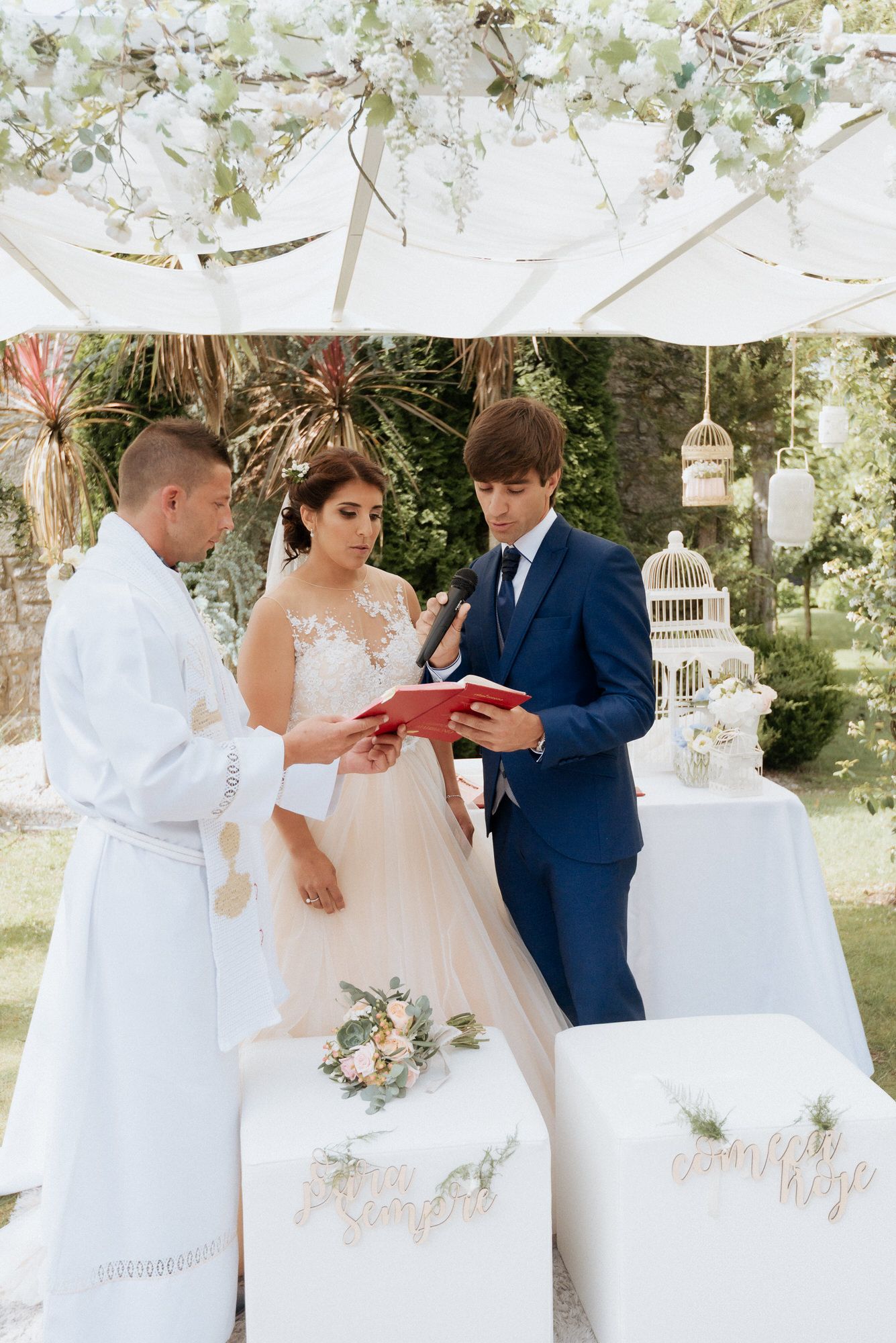 People in formal wedding attire gather for a ceremony under an outdoor canopy adorned with white decorations.