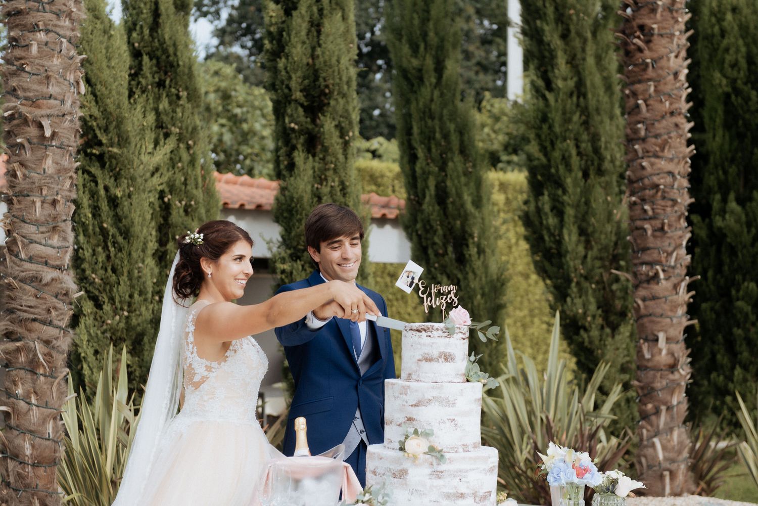 A couple in wedding attire cuts their elegant white marble-effect wedding cake together outdoors among tall cypress trees.