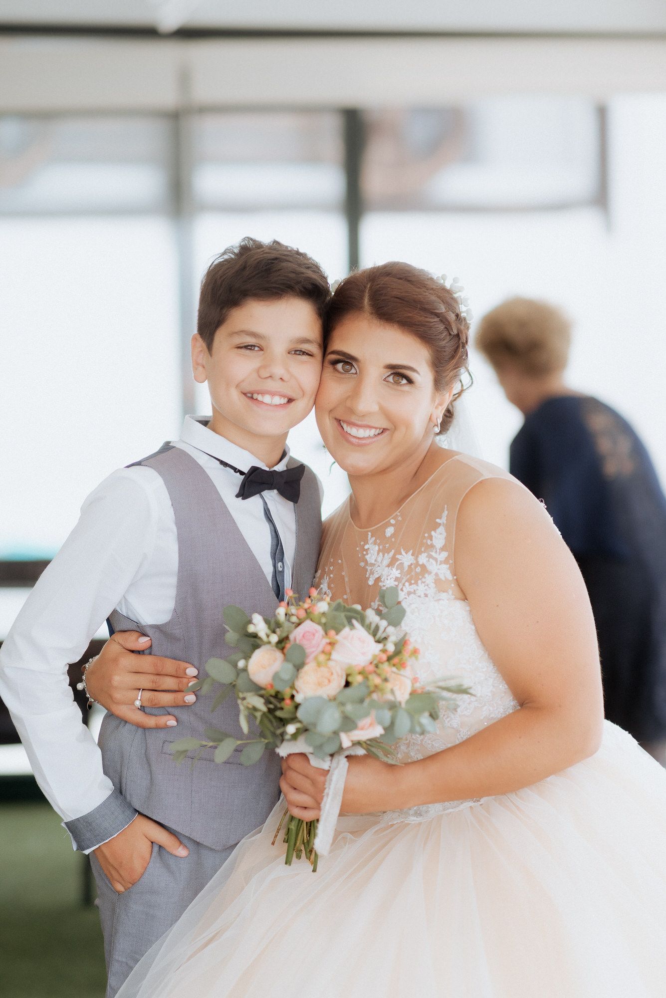 A bride in a white lace gown poses with a young attendant in a gray vest at an indoor wedding venue.