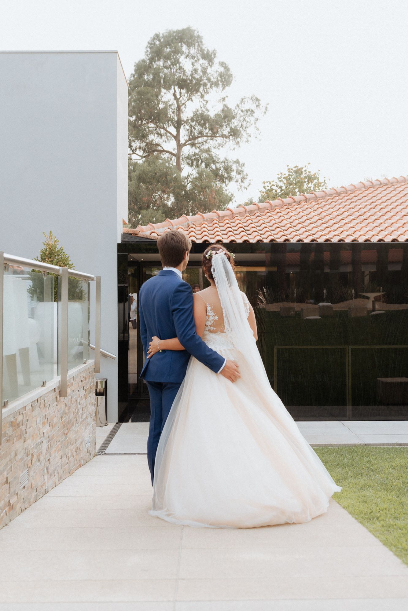 A sequence of five connected photos shows a couple in wedding attire walking down a light-colored walkway with Spanish tile roofing.