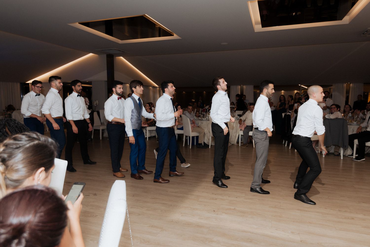 Several people in white shirts and dark pants dancing in formation on a wooden dance floor at an indoor event.