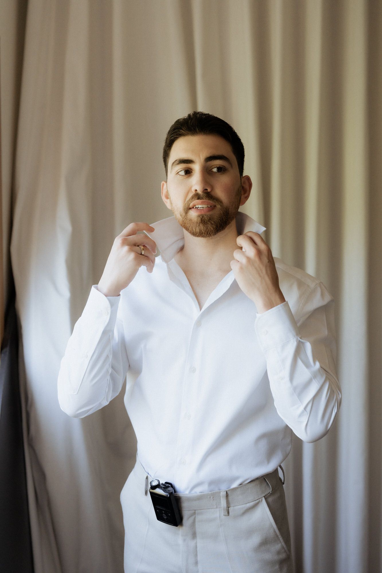 Person wearing a crisp white dress shirt and light colored pants adjusts their collar against a curtained background.