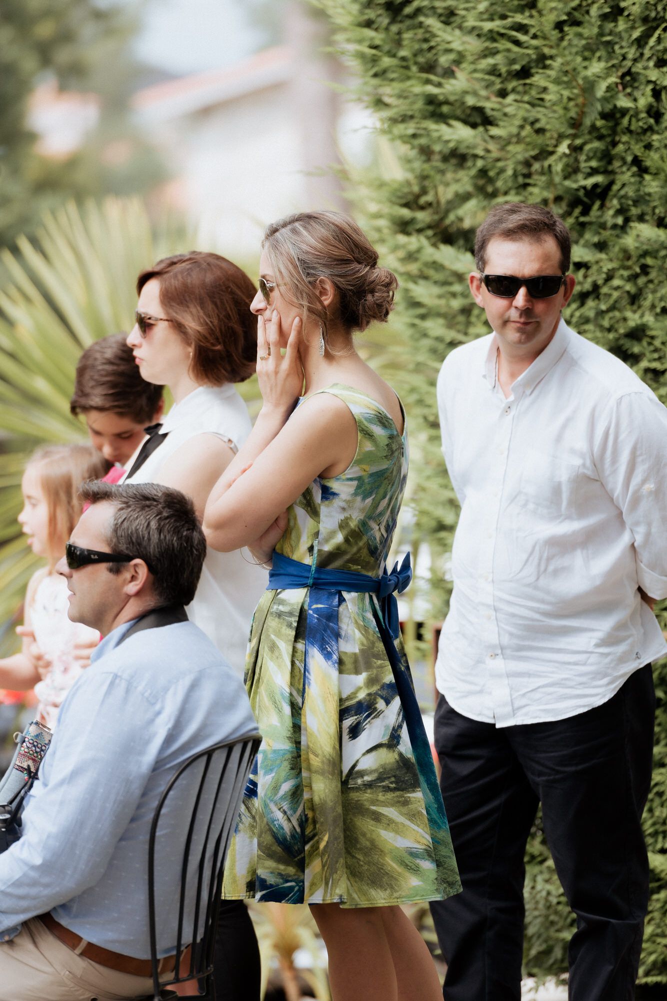 Several guests mingle at an outdoor garden party wearing light summer attire and sunglasses.