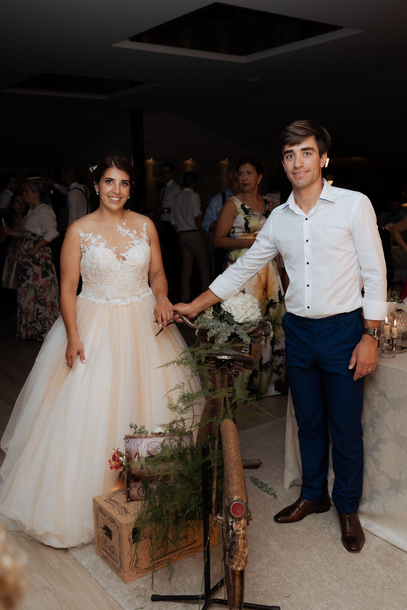 A wedding couple stands together at a reception with rustic wooden decor and greenery arrangements.