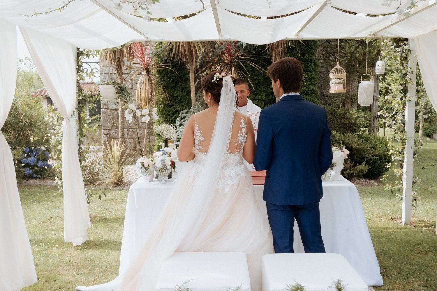 A couple stands under a romantic white draped canopy decorated with hanging lanterns during an outdoor wedding ceremony.
