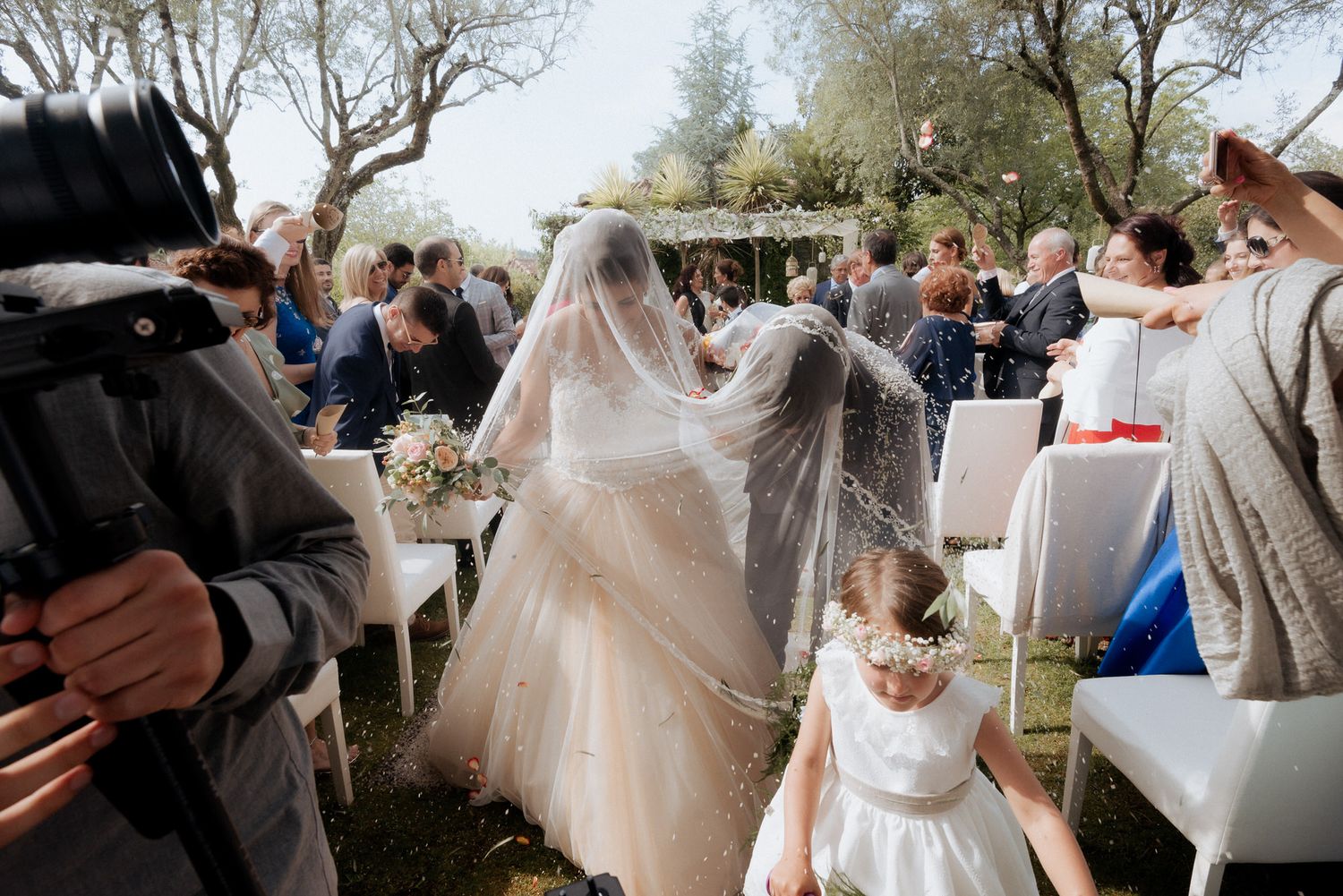 Wedding ceremony guests gather outdoors under bare trees as someone in a flowing white veil walks down the aisle.
