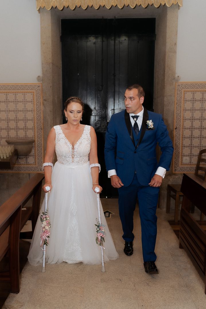 A couple at their wedding ceremony seated in a church, with the bride in white and groom in navy blue suit.