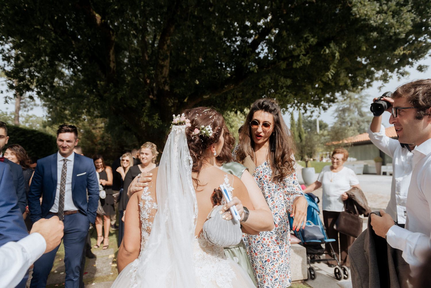 Wedding guests gather outdoors under trees to congratulate and celebrate with the bride at a sunny outdoor ceremony.
