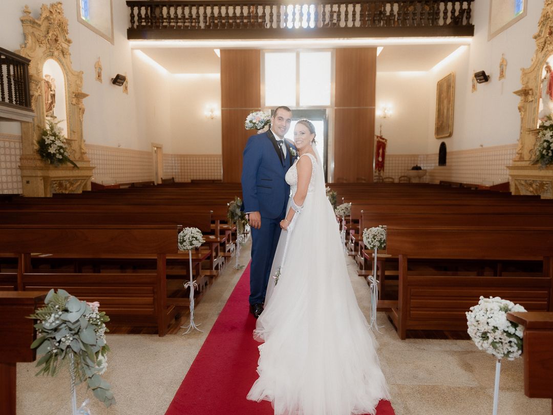 A couple stands together at the altar of an elegant church sanctuary with wooden pews and white floral arrangements.