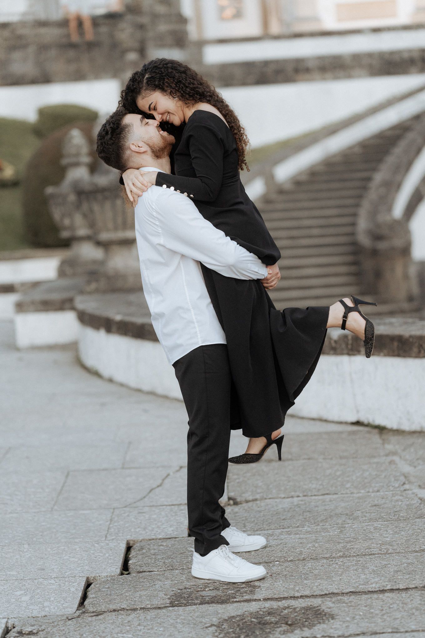 A couple embraces on stone steps, one wearing white and black casual wear, the other in a black dress and heels.