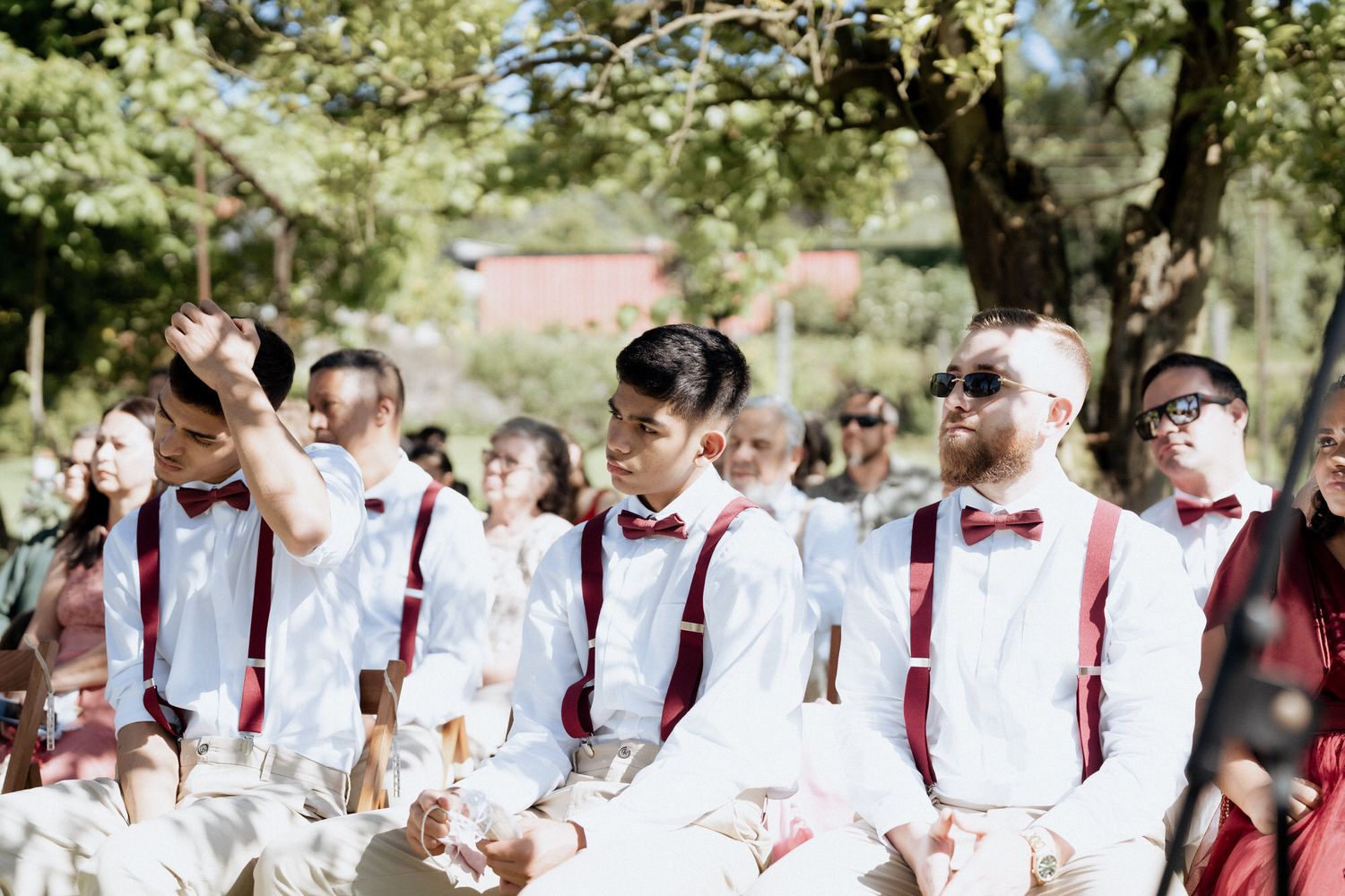 Wedding party members in burgundy bow ties and suspenders stand together in a line at an outdoor ceremony.