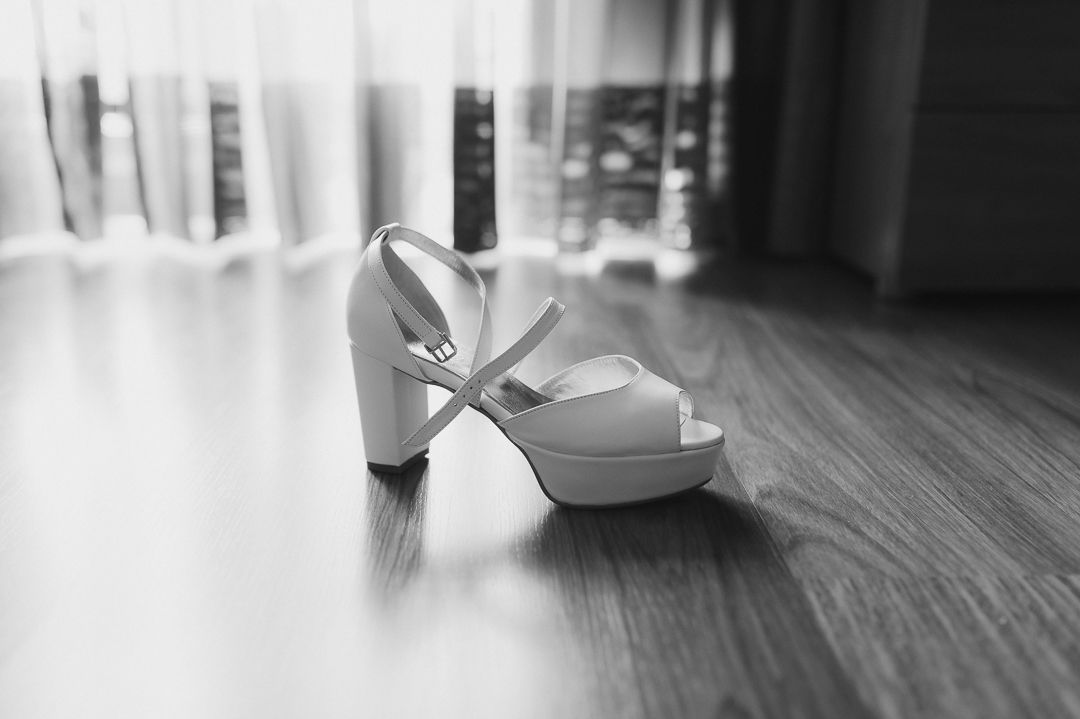 White peep-toe bridal shoe with bow detail sits on hardwood floor in black and white photography.