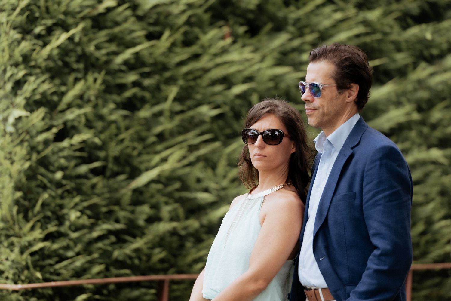 Stylish couple wearing sunglasses poses together outdoors against lush green hedge background.