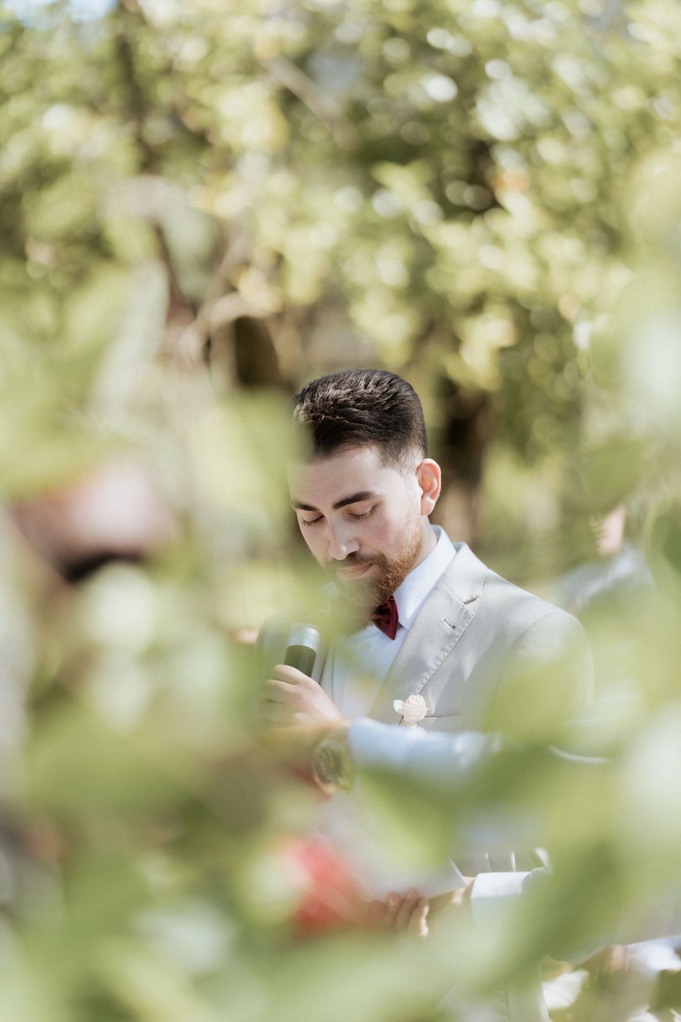A suited figure is photographed through green foliage, gazing downward with a contemplative expression.
