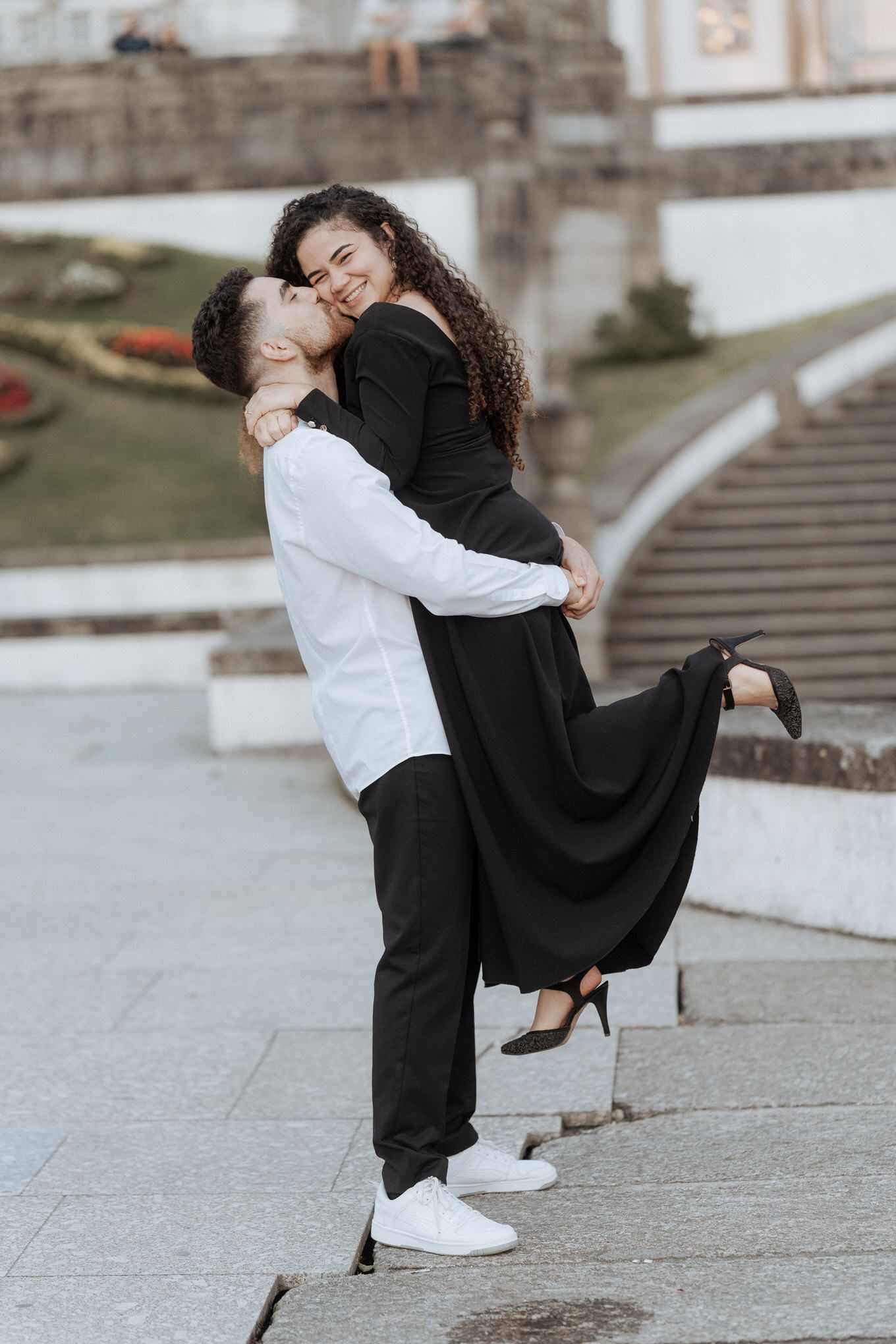 A couple shares a joyful embrace on stone steps, with the person in black wearing a flowing dress and heels.