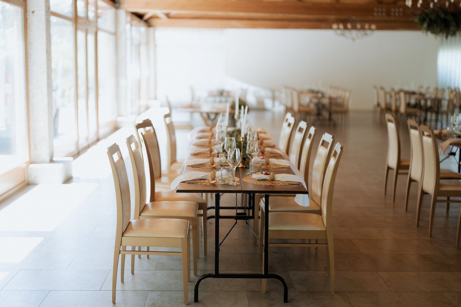 Long rustic dining tables with white chairs set for an elegant event in a bright sunlit venue space.
