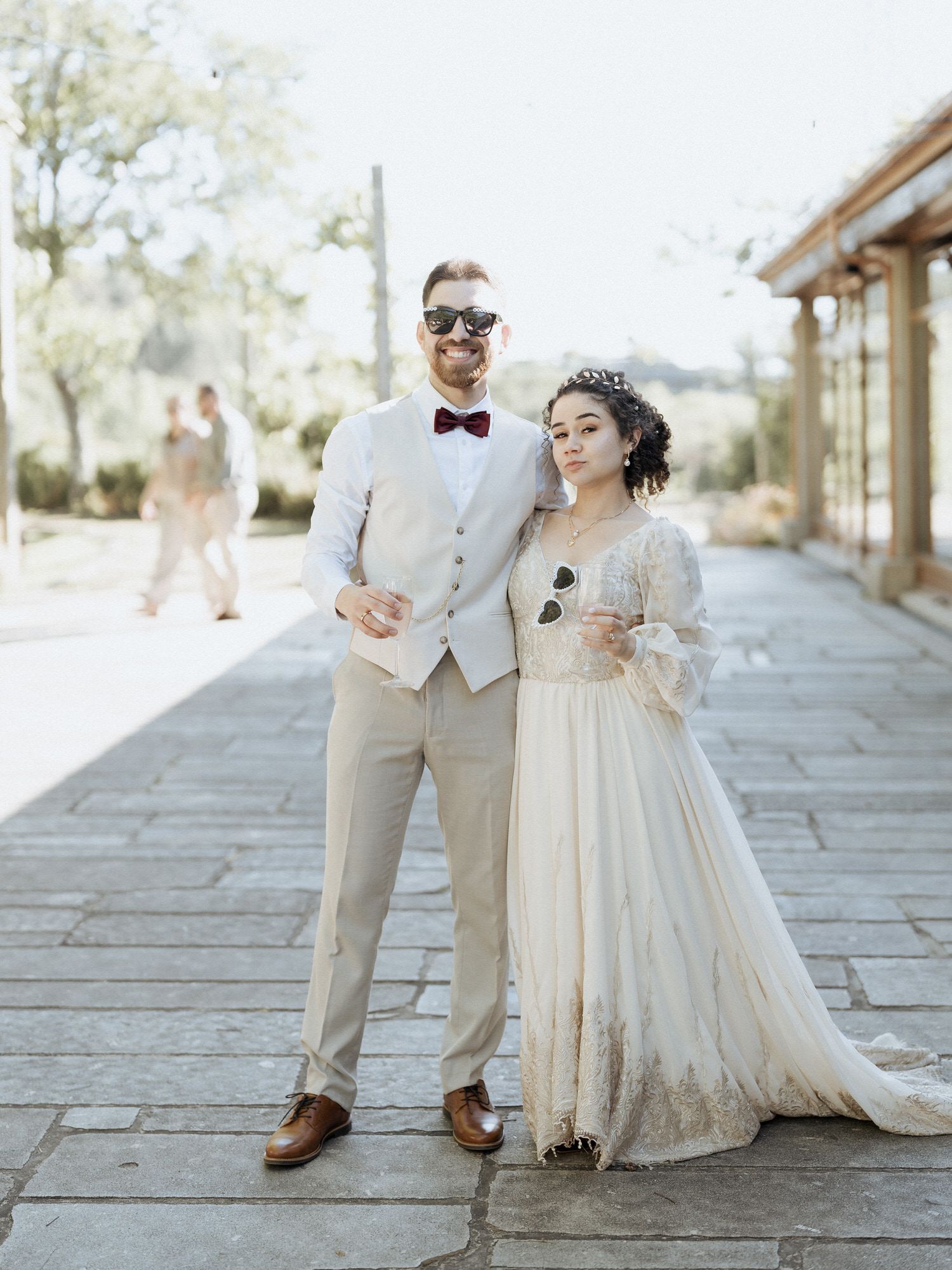 A bride in a vintage lace wedding dress and groom in light suit pose on a stone pathway at a sunlit outdoor venue.