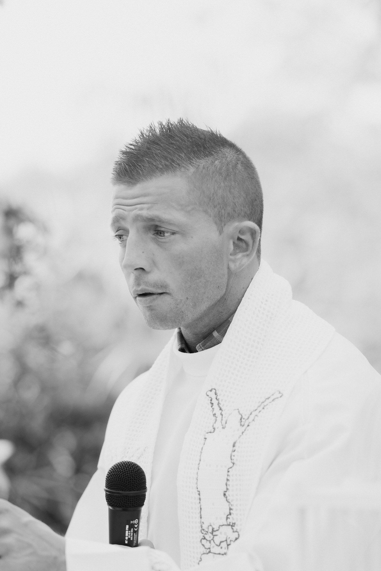 A person speaking into a microphone while wearing a white garment with embroidered details in a black and white image.