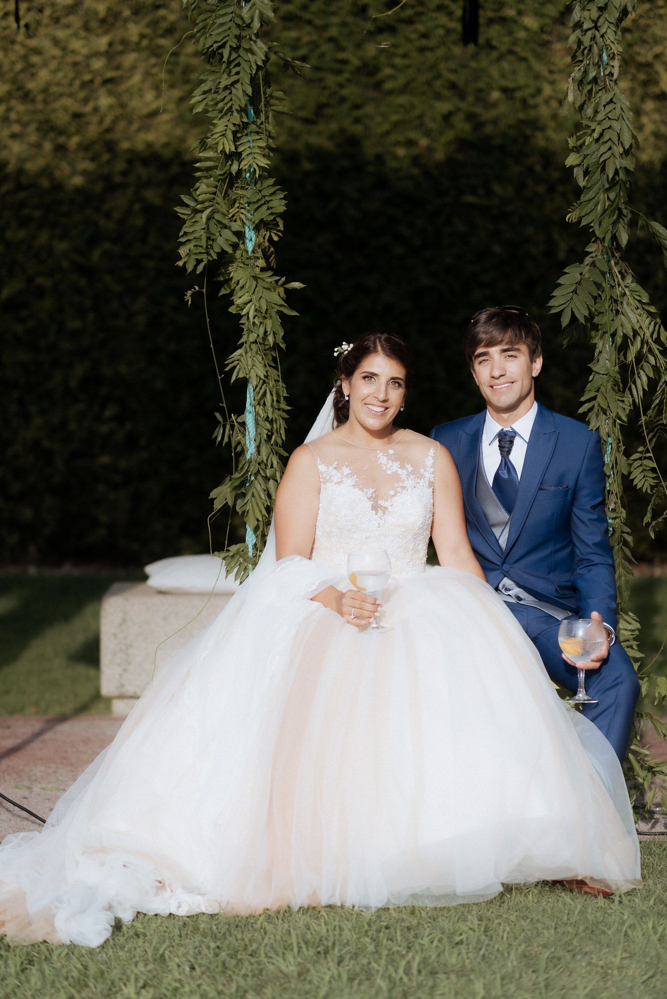 A bride in a flowing white tulle gown and groom in a blue suit sit together on a garden swing adorned with greenery.