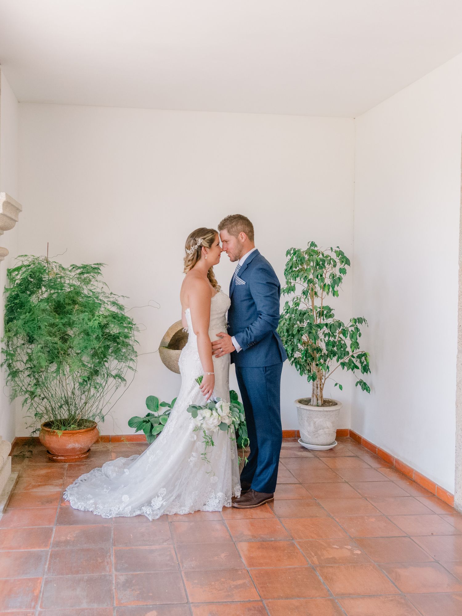 Casal de recém-casados compartilhando um momento íntimo em uma sala branca com vasos de plantas e piso de terracota.