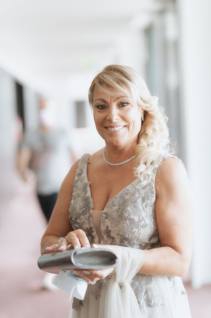 A person in a light gray formal dress with beaded details and a pearl necklace smiles while holding a clutch purse.