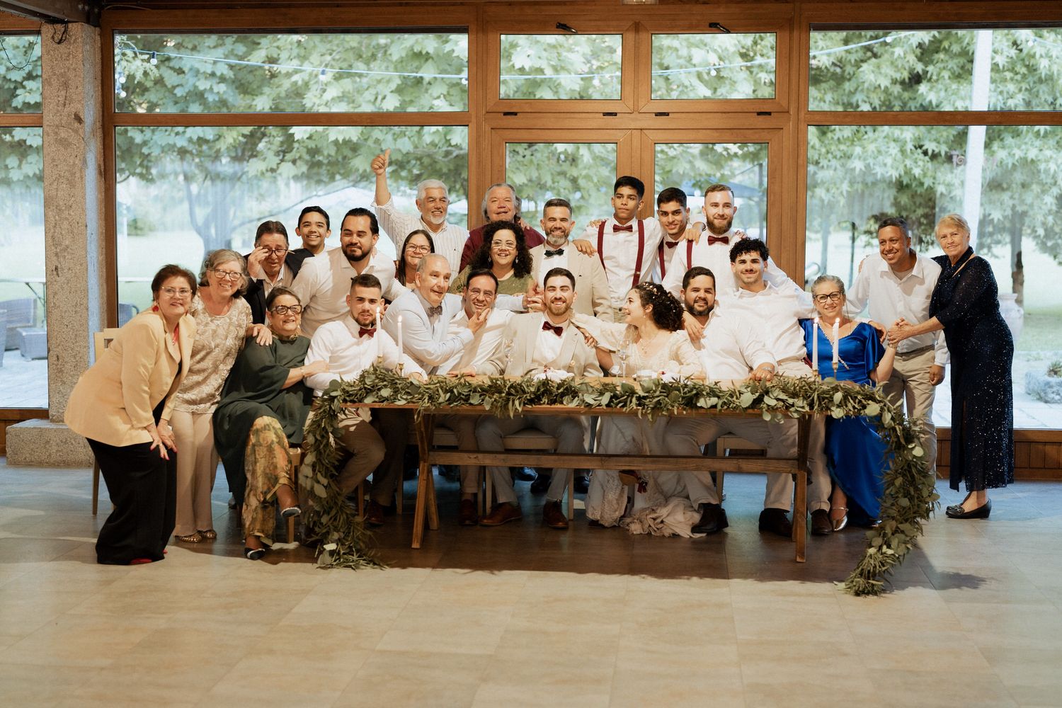 Large group poses together behind a decorated rustic wooden table at an indoor-outdoor wedding venue.