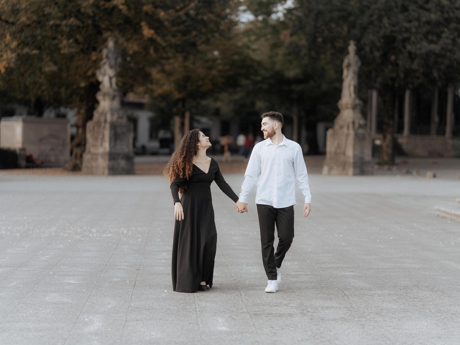 Um casal de mãos dadas enquanto caminham juntos em um pátio elegante com esculturas de pedra e árvores ao fundo.